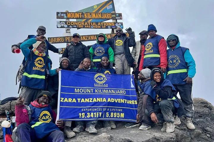 Guides and porters holding a banner at the Uhuru Peak sign on a Kilimanjaro climbing tour by the Marangu Route, Tanzania